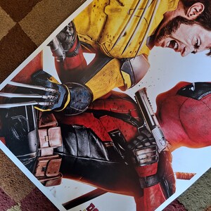 以下が含まれることがあります： デッドプールとウルヴァリンが戦っている映画のポスター。デッドプールは、特徴的な赤と黒の衣装を着て、手前にいます。ウルヴァリンは、黄色と黒の衣装を着て、奥にいます。ポスターには、黄色と黒で「DEADPOOL & WOLVERINE」と書かれています。