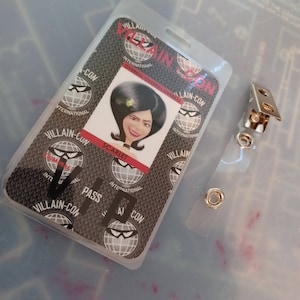 May include: A white plastic badge with a black and white design. The badge features a cartoon image of a woman with black hair and a green bow. The text on the badge reads "VILLAIN-CON INTERNATIONAL OVERKILL OVERKILL OVERKILL" and "SCARLET".