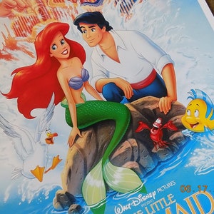 May include: A poster for the Disney movie The Little Mermaid featuring Ariel, a red-haired mermaid, and Prince Eric, a human prince. Ariel is sitting on a rock with her tail in the water, while Eric is standing behind her. The poster also features a seagull, a crab, and a yellow fish.