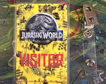 May include: A yellow and black Jurassic World visitor badge with a dinosaur graphic and the text "Jurassic World" and "Visitor". The badge is in a clear plastic holder with a clip.