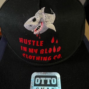 Può includere: Cappellino da baseball nero con un grafico di squalo bianco e ricamo rosso che recita "Hustle in my blood Clothing Co.". Il cappello ha un'etichetta argentata che recita "Otto S.N.A.P. One Size Fits Most."