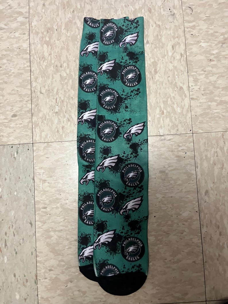 K&ouml;nnte beinhalten: Ein Paar gr&uuml;n-schwarze Socken mit einem sich wiederholenden Muster des Philadelphia Eagles-Logos. Das Logo ist ein wei&szlig;er Adler mit schwarzem Umriss und den Worten "Philadelphia Eagles" in einem schwarzen Kreis.