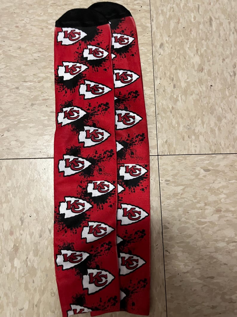 K&ouml;nnte beinhalten: Ein Paar rot-schwarze Socken mit einem wei&szlig;en Kansas City Chiefs-Logo, das sich auf dem Stoff wiederholt. Die Socken haben ein Spr&uuml;hfarben-Design.