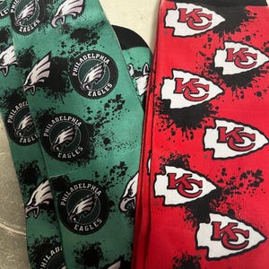 K&ouml;nnte beinhalten: Ein Paar Socken, eine gr&uuml;n mit dem Philadelphia Eagles Logo und die andere rot mit dem Kansas City Chiefs Logo. Beide Socken haben ein Spritzlack-Design.