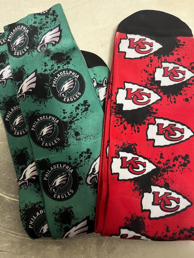 K&ouml;nnte beinhalten: Ein Paar Socken, eine gr&uuml;n mit dem Philadelphia Eagles Logo und die andere rot mit dem Kansas City Chiefs Logo. Beide Socken haben ein Spritzlack-Design.