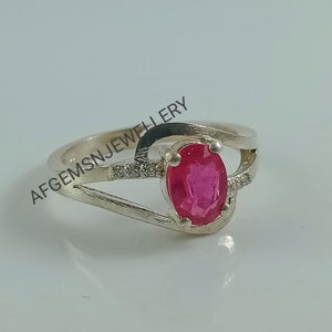 Puede incluir: Un anillo de plata con una piedra ovalada rosa engastada en un dise&ntilde;o delicado y curvo. El anillo tiene una peque&ntilde;a fila de piedras transparentes a cada lado de la piedra principal. AFGEMSNJEWELLERY est&aacute; impreso en la banda.
