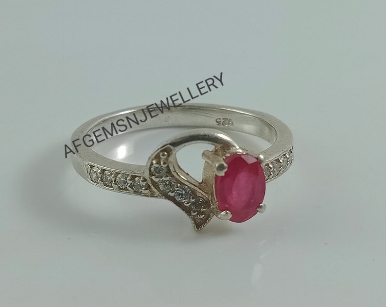 May include: A silver ring with a red oval gemstone and small clear stones set in the band. The ring is marked "925" on the inside.