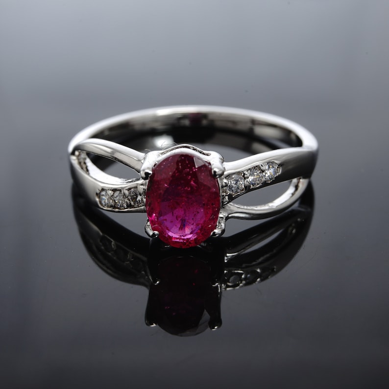 Pode incluir: Um anel de prata com uma grande pedra de rubi oval e pedras menores transparentes engastadas na banda.