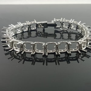 Op de afbeelding: Een zilverkleurige armband met vierkante zettingen ontworpen om edelstenen te bevatten. De armband wordt getoond op een reflecterend oppervlak, wat het ingewikkelde ontwerp en de individuele zettingen voor stenen benadrukt. De sluiting is zichtbaar.