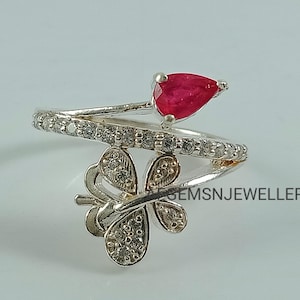 K&ouml;nnte beinhalten: Ein silberner Ring mit einem roten Edelstein und einem Schmetterlingsdesign. Der Ring ist mit kleinen, klaren Steinen besetzt.