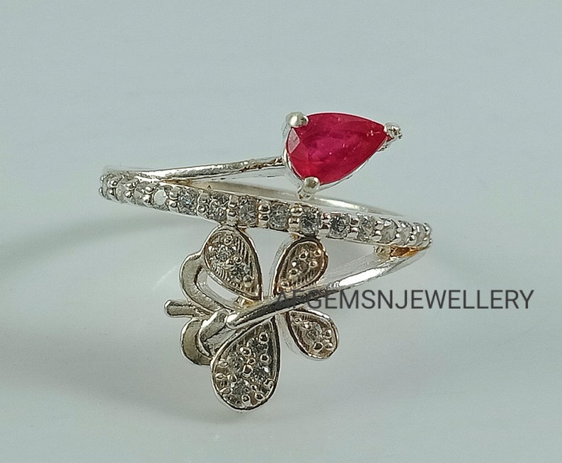 K&ouml;nnte beinhalten: Ein silberner Ring mit einem roten Edelstein und einem Schmetterlingsdesign. Der Ring ist mit kleinen, klaren Steinen besetzt.