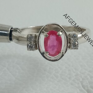 K&ouml;nnte beinhalten: Ein silberner Ring mit einem rosafarbenen ovalen Edelstein, der von einem silbernen Rahmen umgeben ist und auf beiden Seiten zwei kleinere, klare Edelsteine hat. Der Ring liegt auf einer wei&szlig;en Oberfl&auml;che. Im Hintergrund ist der Text 'AFGEMSN JEWELLERY' zu sehen.