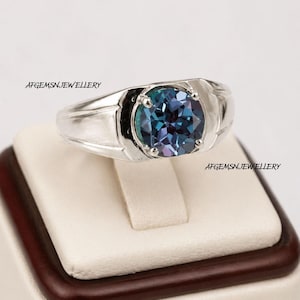 May include: A silver-toned ring featuring a round, faceted, color-changing gemstone in shades of blue and purple. The band has a smooth, polished finish. The text AFGEMSNJEWELLERY is visible twice on the image near the ring.