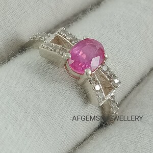 May include: A silver ring featuring a pink oval gemstone surrounded by smaller white gemstones. The ring is displayed on a white fabric background. The text "AFGEMS JEWELLERY" is visible in the bottom right corner of the image.