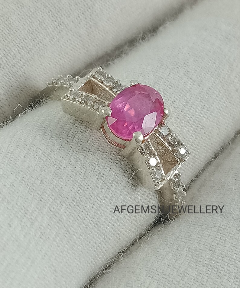 May include: A silver ring featuring a pink oval gemstone surrounded by smaller white gemstones. The ring is displayed on a white fabric background. The text "AFGEMS JEWELLERY" is visible in the bottom right corner of the image.