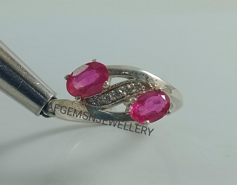 May include: A silver ring with two oval-shaped pink gemstones and a row of smaller white gemstones. The ring is engraved with the text "AFGEMS JEWELLERY".