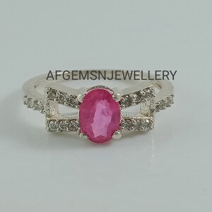 May include: A silver ring featuring a pink oval gemstone set within a delicate design of smaller white gemstones. The ring has two bands that cross over each other. The text "AFGEMSNJEWELLERY" is visible above the ring.