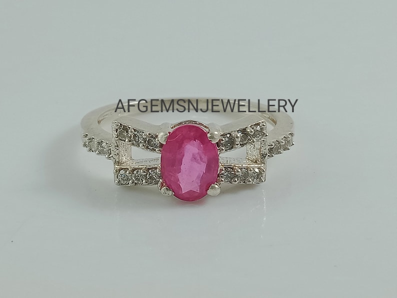 May include: A silver ring featuring a pink oval gemstone set within a delicate design of smaller white gemstones. The ring has two bands that cross over each other. The text "AFGEMSNJEWELLERY" is visible above the ring.