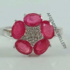 Puede incluir: Un anillo de plata con un grupo de seis piedras preciosas ovaladas de color rosa engastadas en un diseño floral. El anillo está sobre un fondo blanco.