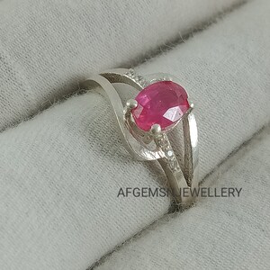 Puede incluir: Un anillo de plata con una piedra ovalada rosa y piedras m&aacute;s peque&ntilde;as transparentes engastadas en la banda. AFGEMS JEWELLERY est&aacute; impreso en la superficie debajo del anillo.