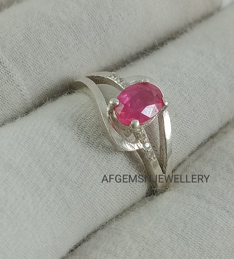 Puede incluir: Un anillo de plata con una piedra ovalada rosa y piedras m&aacute;s peque&ntilde;as transparentes engastadas en la banda. AFGEMS JEWELLERY est&aacute; impreso en la superficie debajo del anillo.