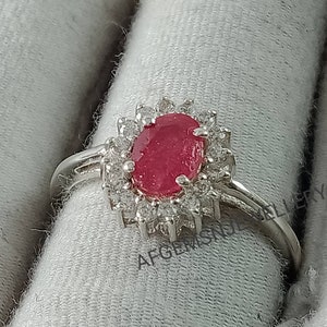 Pu&ograve; includere: Un anello d'argento con un grande rubino ovale circondato da pietre bianche pi&ugrave; piccole. L'anello &egrave; su uno sfondo di tessuto bianco.