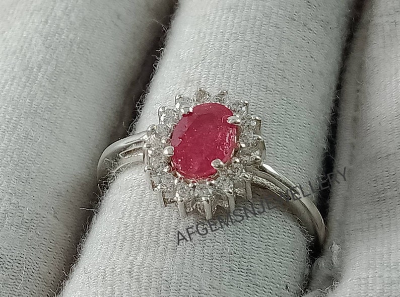 Pu&ograve; includere: Un anello d'argento con un grande rubino ovale circondato da pietre bianche pi&ugrave; piccole. L'anello &egrave; su uno sfondo di tessuto bianco.