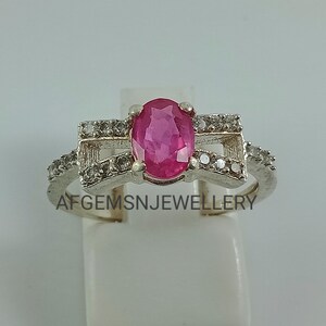 May include: A silver ring featuring a pink oval gemstone surrounded by smaller clear gemstones. The ring has a delicate design with two curved bands that meet in the middle.