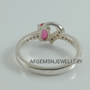 May include: A silver ring with a pink oval gemstone and smaller clear gemstones set in a heart-shaped design. The ring is inscribed with the text "AFGEMSNJEWELLERY".