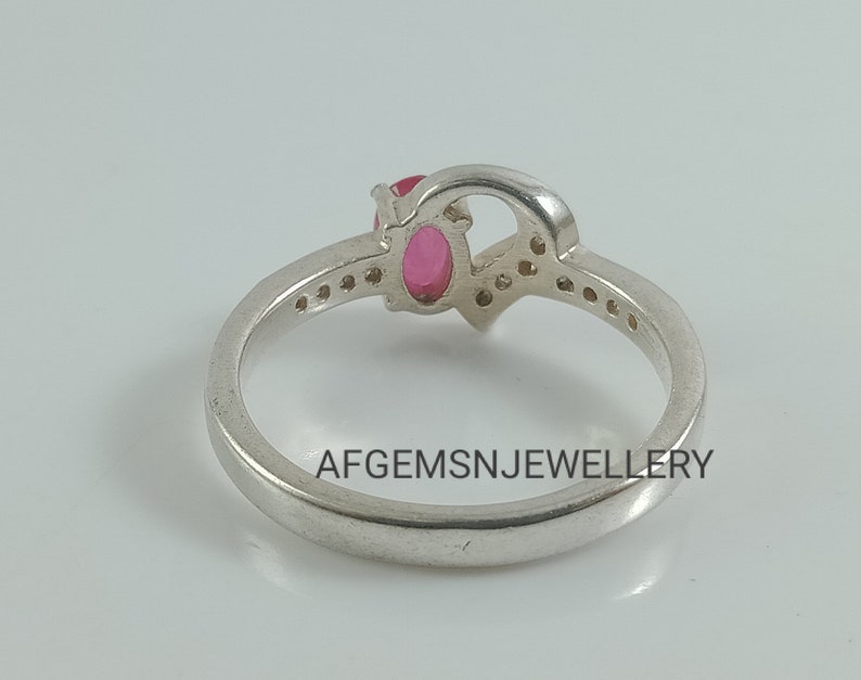 May include: A silver ring with a pink oval gemstone and smaller clear gemstones set in a heart-shaped design. The ring is inscribed with the text "AFGEMSNJEWELLERY".