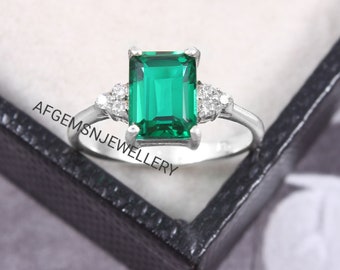 May include: A silver ring with a large emerald green gemstone and smaller clear gemstones on the band. The ring is in a black box with the text "AFGEMSNJEWELLERY".