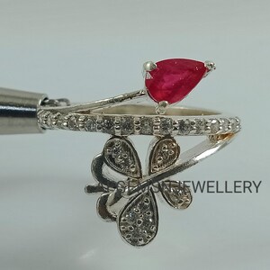K&ouml;nnte beinhalten: Ein silberner Ring mit einem roten birnenf&ouml;rmigen Edelstein und einem Schmetterlingsdesign mit klaren Steinen. Der Ring liegt auf einem wei&szlig;en Hintergrund.