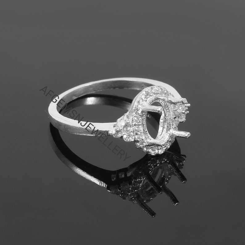 May include: A silver ring with a prong setting for an oval-shaped gemstone. The ring is decorated with small, clear stones around the setting.
