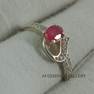 May include: A silver ring with a heart-shaped design and a red ruby gemstone. The ring is set with small clear gemstones around the ruby and the band.