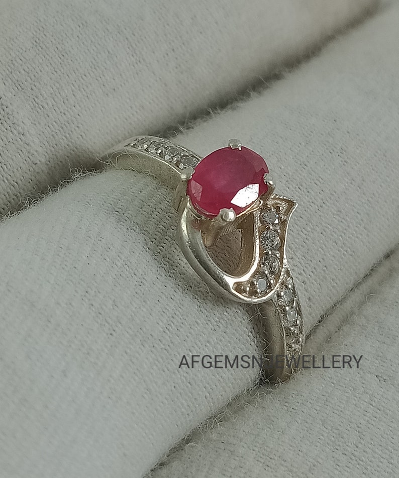 May include: A silver ring with a heart-shaped design and a red ruby gemstone. The ring is set with small clear gemstones around the ruby and the band.