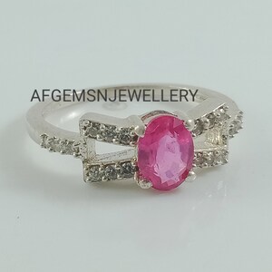 May include: A silver ring featuring a pink oval gemstone set within a delicate design of smaller white gemstones. The ring has two bands that cross over each other. The text "AFGEMSNJEWELLERY" is visible on the band.