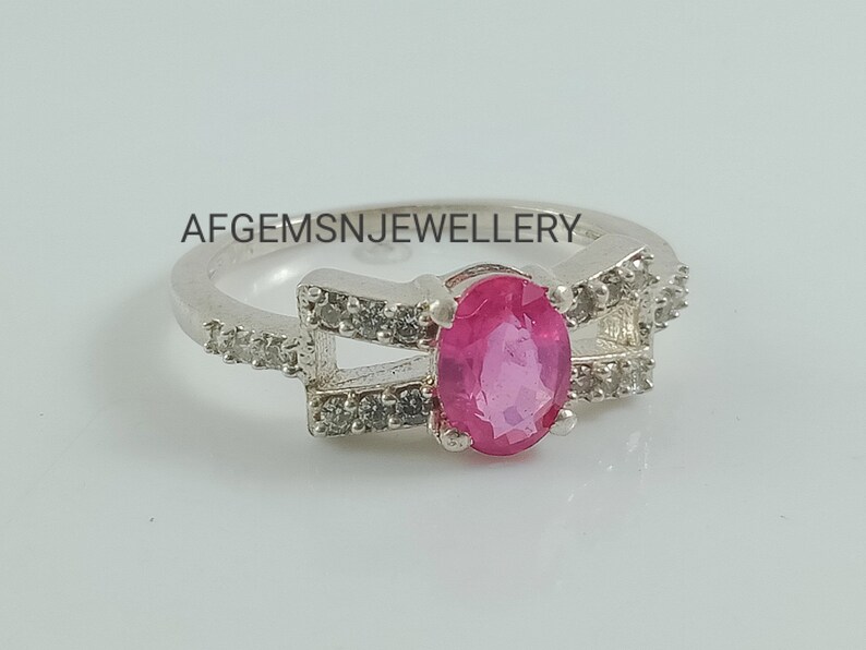 May include: A silver ring featuring a pink oval gemstone set within a delicate design of smaller white gemstones. The ring has two bands that cross over each other. The text "AFGEMSNJEWELLERY" is visible on the band.