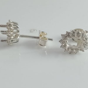 May include: A pair of silver stud earrings. One earring is fully assembled with a central oval setting surrounded by small, round stones. The other earring is a setting with prongs for holding a stone, and a post.