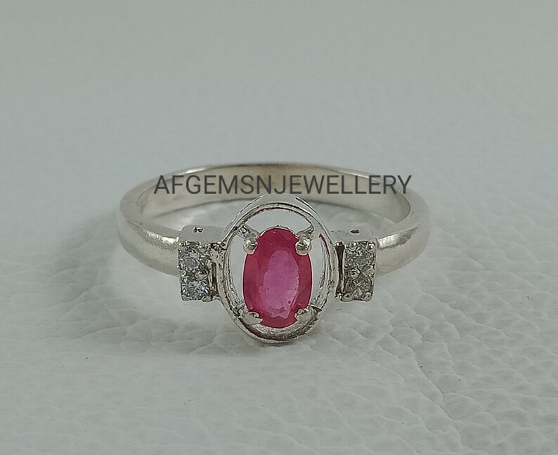 K&ouml;nnte beinhalten: Ein silberner Ring mit einem rosafarbenen ovalen Edelstein, der von einem silbernen Rahmen umgeben ist und auf beiden Seiten zwei kleinere klare Edelsteine hat. Der Ring liegt auf einer wei&szlig;en Oberfl&auml;che. Auf der Ringschiene ist 'AFGEMSNJEWELLERY' eingraviert.