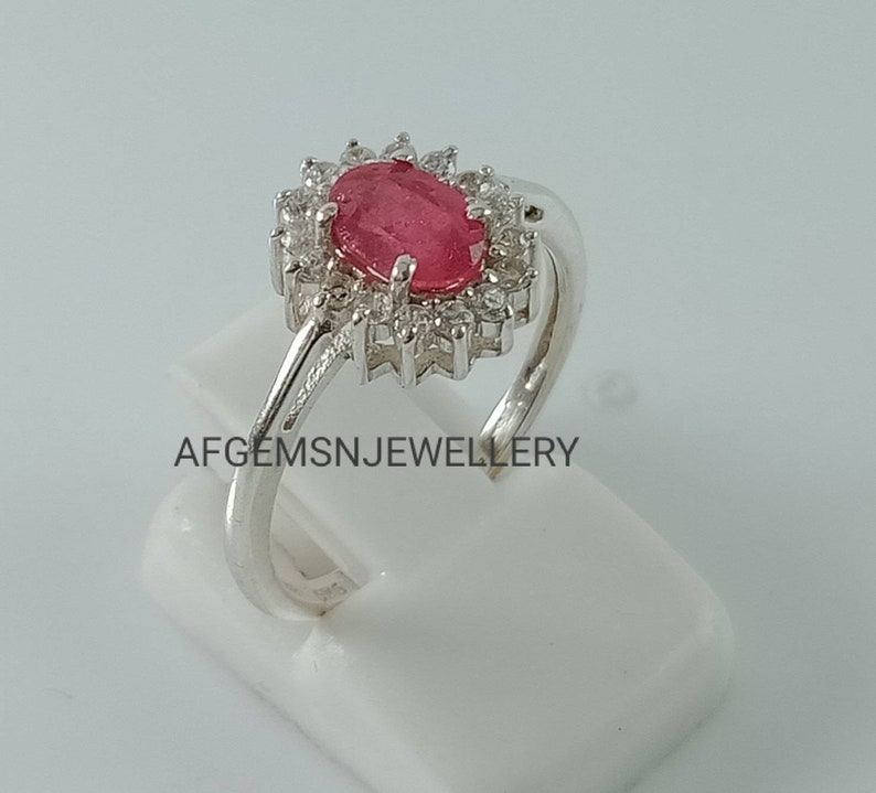 Pu&ograve; includere: Un anello d'argento con una pietra ovale rosa circondata da pietre bianche pi&ugrave; piccole. L'anello &egrave; su un espositore bianco. Il testo 'AFGEMSNJEWELLERY' &egrave; stampato sull'espositore.