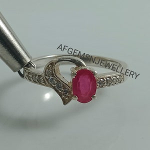 May include: A silver ring with a heart-shaped design and a red oval gemstone. The ring is set with small white gemstones around the band. The text "AFGEMS N JEWELLERY" is visible on the band.