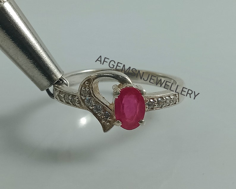 May include: A silver ring with a heart-shaped design and a red oval gemstone. The ring is set with small white gemstones around the band. The text "AFGEMS N JEWELLERY" is visible on the band.