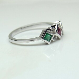 May include: A silver band ring with three gemstones set in a diamond shape. The gemstones are a green emerald, a red ruby, and a clear diamond.