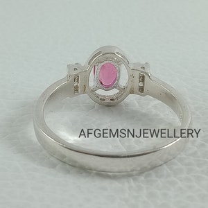 K&ouml;nnte beinhalten: Ein silberner Ring mit einem rosafarbenen ovalen Edelstein, der in einer Halo-Fassung sitzt. Der Ring hat kleine klare Edelsteine auf jeder Seite des Hauptsteins. Die Band ist glatt und einfach. AFGEMSNJEWELLERY ist auf der R&uuml;ckseite der Band aufgedruckt.