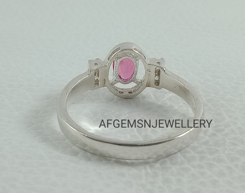 K&ouml;nnte beinhalten: Ein silberner Ring mit einem rosafarbenen ovalen Edelstein, der in einer Halo-Fassung sitzt. Der Ring hat kleine klare Edelsteine auf jeder Seite des Hauptsteins. Die Band ist glatt und einfach. AFGEMSNJEWELLERY ist auf der R&uuml;ckseite der Band aufgedruckt.