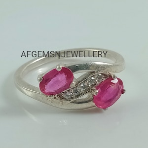 May include: A silver ring with two pink oval gemstones and smaller clear gemstones set in a twisted band. The ring is on a white background. AFGEMSNJEWELLERY is written in the background.