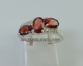 May include: A silver ring with three red oval gemstones and small white stones around the band. The ring is on a white display stand.  AFGEMSNJEWELLERY