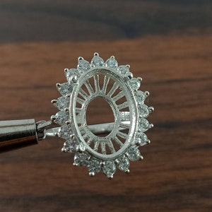 May include: A silver ring setting with an oval centre and a halo of small, round, clear gemstones. The setting is designed to hold a gemstone and is displayed against a dark wood background.
