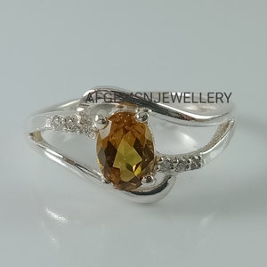 Può includere: Un anello d'argento con una grande gemma ovale giallo dorato. L'anello presenta un design a spirale con piccole pietre chiare. Il testo "AFGESNJEWELLERY" è visibile sull'anello.