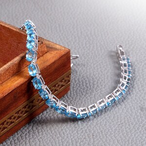 May include: A silver bracelet with a row of oval-shaped blue gemstones. The bracelet is lying on a wooden box with intricate carvings.